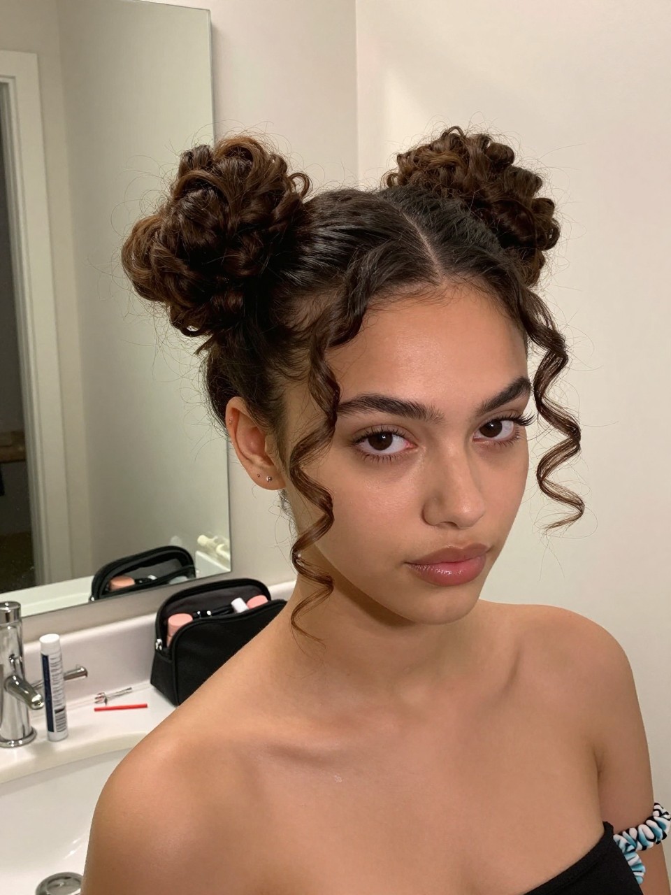 Image d'illustration : Photo d'une femme portant deux chignons bouclés souples de type 4A avec une raie au milieu nette, des pointes de boucles lâches autour de chaque chignon et deux mèches définies encadrant le visage, vue de trois quarts avant à côté d'un miroir de salle de bain, éclairage intérieur doux, contenant un petit détail du quotidien comme des épingles à cheveux et du gel pour boucles sur le comptoir, une trousse de maquillage à proximité et deux chouchous de rechange à son poignet, moment réaliste de préparation, qualité photo iPhone de dernière génération.