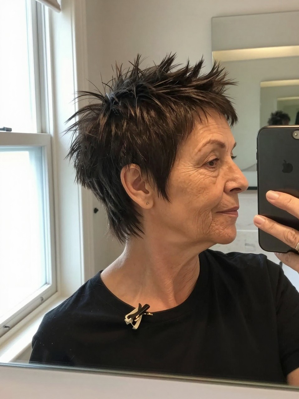 Photo d'une femme âgée portant une coupe pixie hérissée, selfie de profil dans un miroir montrant la texture relevée au niveau de la couronne, prise devant un miroir de salle de bain avec lumière naturelle, avec une pince à cheveux accrochée à son col, qualité photo iPhone.