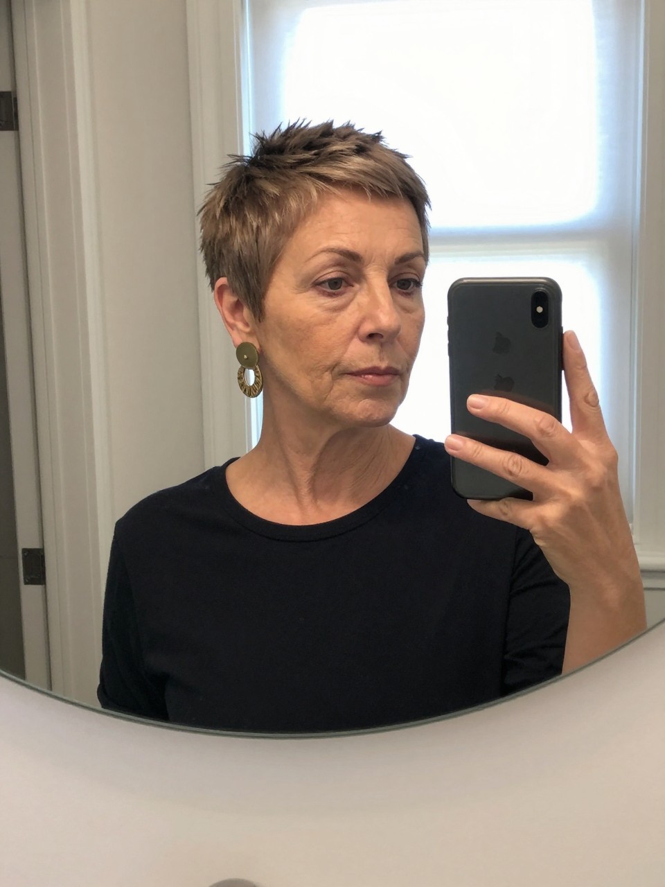 Photo d'une femme d'un certain âge portant une coupe pixie courte et texturée, selfie pris devant un miroir de face montrant le cadrage serré, le décor est un miroir de coiffeuse avec lumière naturelle, contenant une paire de boucles d'oreilles originales visibles, qualité photo iPhone.