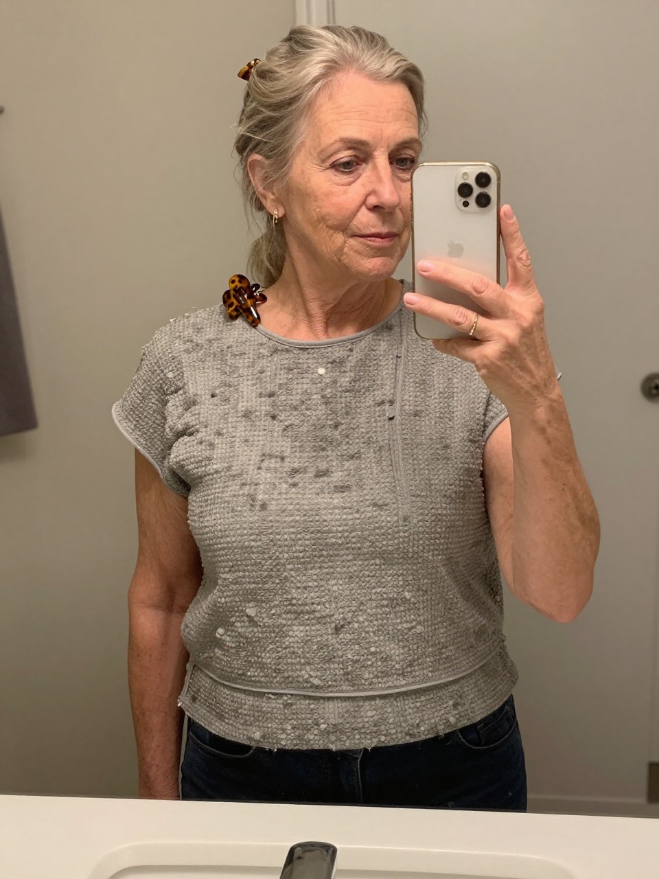 Photo d'une femme âgée portant un crop top texturé, vue de face montrant les différentes couches, le décor est un miroir de salle de bain avec un éclairage intérieur doux, avec une barrette à cheveux en écaille de tortue accrochée à son col, qualité photo iPhone.