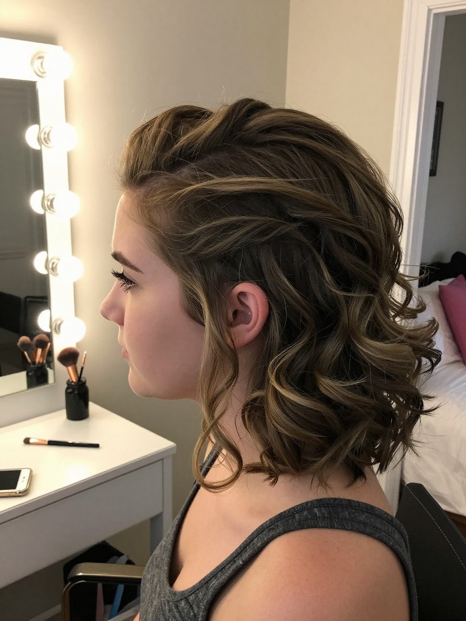 Photo d'une adolescente américaine blanche portant une demi-queue texturée et volumineuse, vue de profil montrant la hauteur au sommet de la tête, décor : une chambre avec une coiffeuse et des ampoules de style hollywoodien, éclairage : lumière intérieure chaude et douce, contenant un pinceau de maquillage posé sur la coiffeuse, qualité photo iPhone.