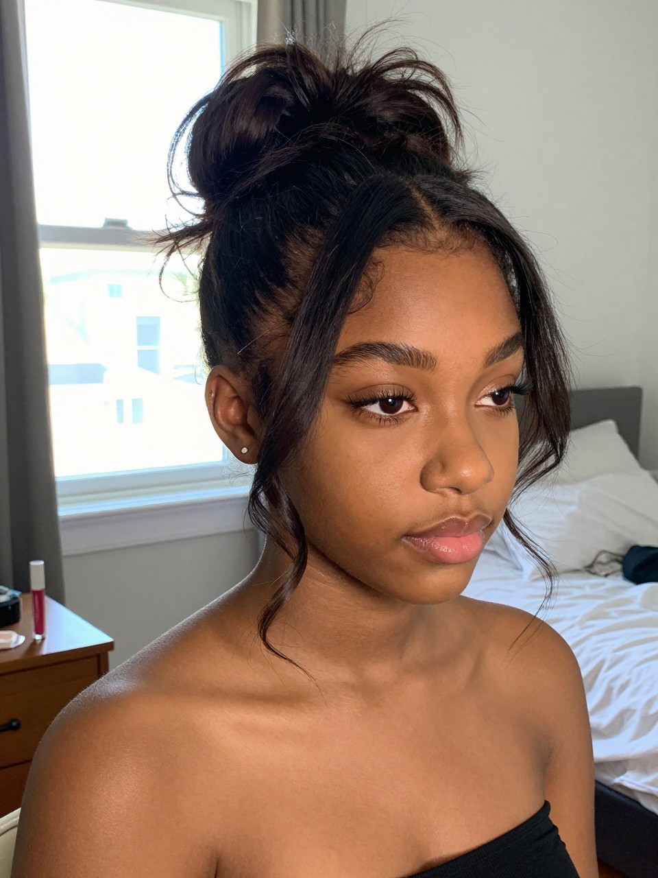 Photo d'une adolescente américaine noire portant une demi-queue de cheval décoiffée avec des mèches encadrant le visage, vue de face en biais montrant les mèches douces autour du visage, décor : une chambre avec lumière naturelle provenant d'une fenêtre, éclairage : lumière douce et vive de la fenêtre, contenant un tube de gloss à lèvres sur une table de nuit, qualité photo iPhone.