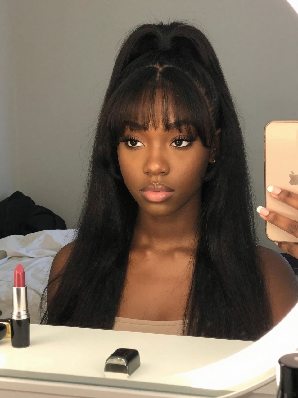 Photo d'une adolescente afro-américaine portant une demi-queue de cheval avec une frange rideau, vue de face montrant la frange encadrant le visage, décor : une chambre avec une coiffeuse et un miroir, éclairage : lumière intérieure douce, contenant un tube de rouge à lèvres sur la coiffeuse, qualité photo iPhone.