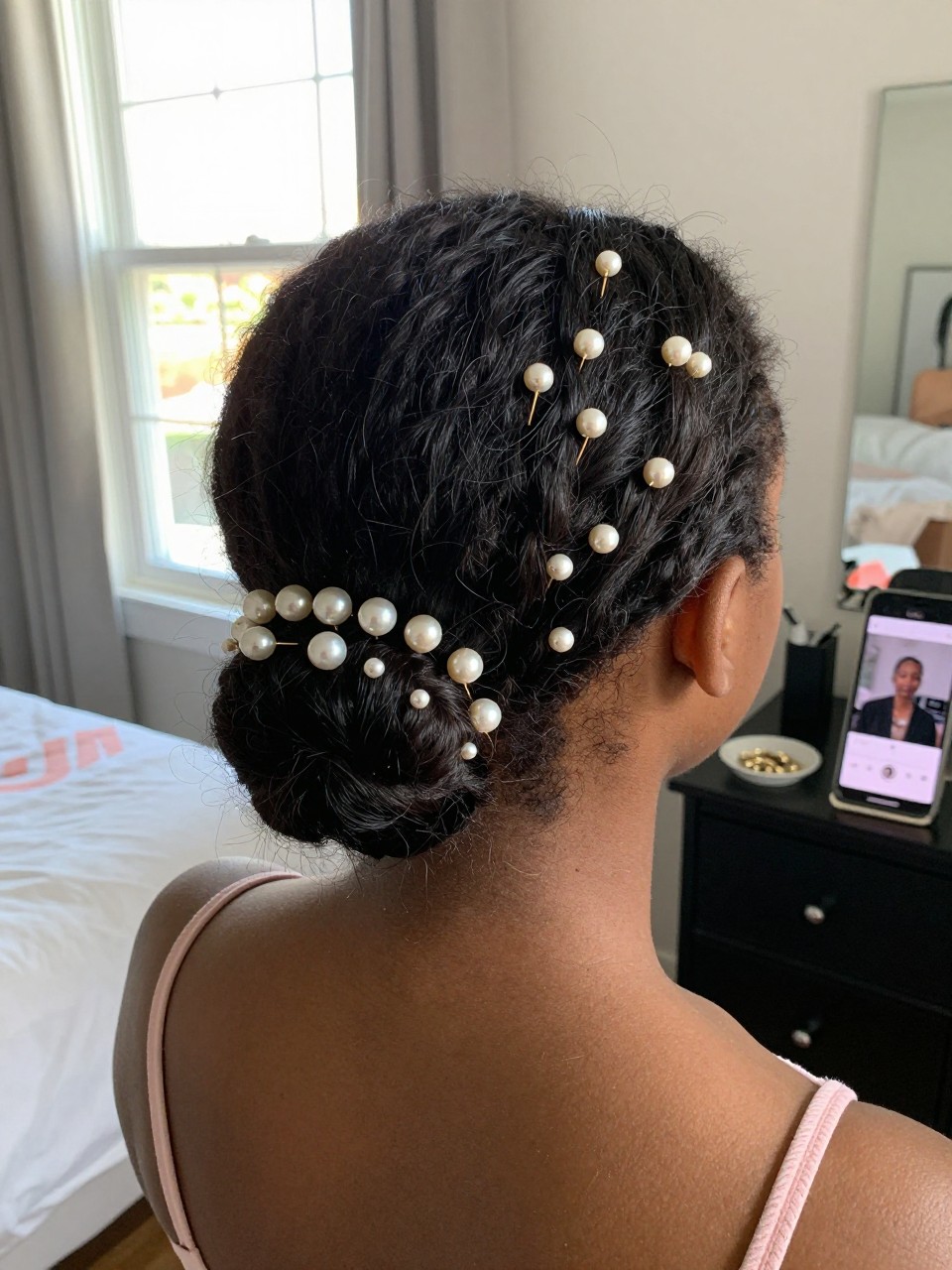 Photo d'une adolescente afro-américaine portant une demi-queue de cheval ornée d'épingles à perles, vue de l'arrière de la tête montrant les épingles à la base, décor : une chambre avec lumière naturelle provenant d'une fenêtre, éclairage : lumière douce et vive de la fenêtre, contenant un petit plat avec des épingles à perles de rechange sur une commode, qualité photo iPhone.