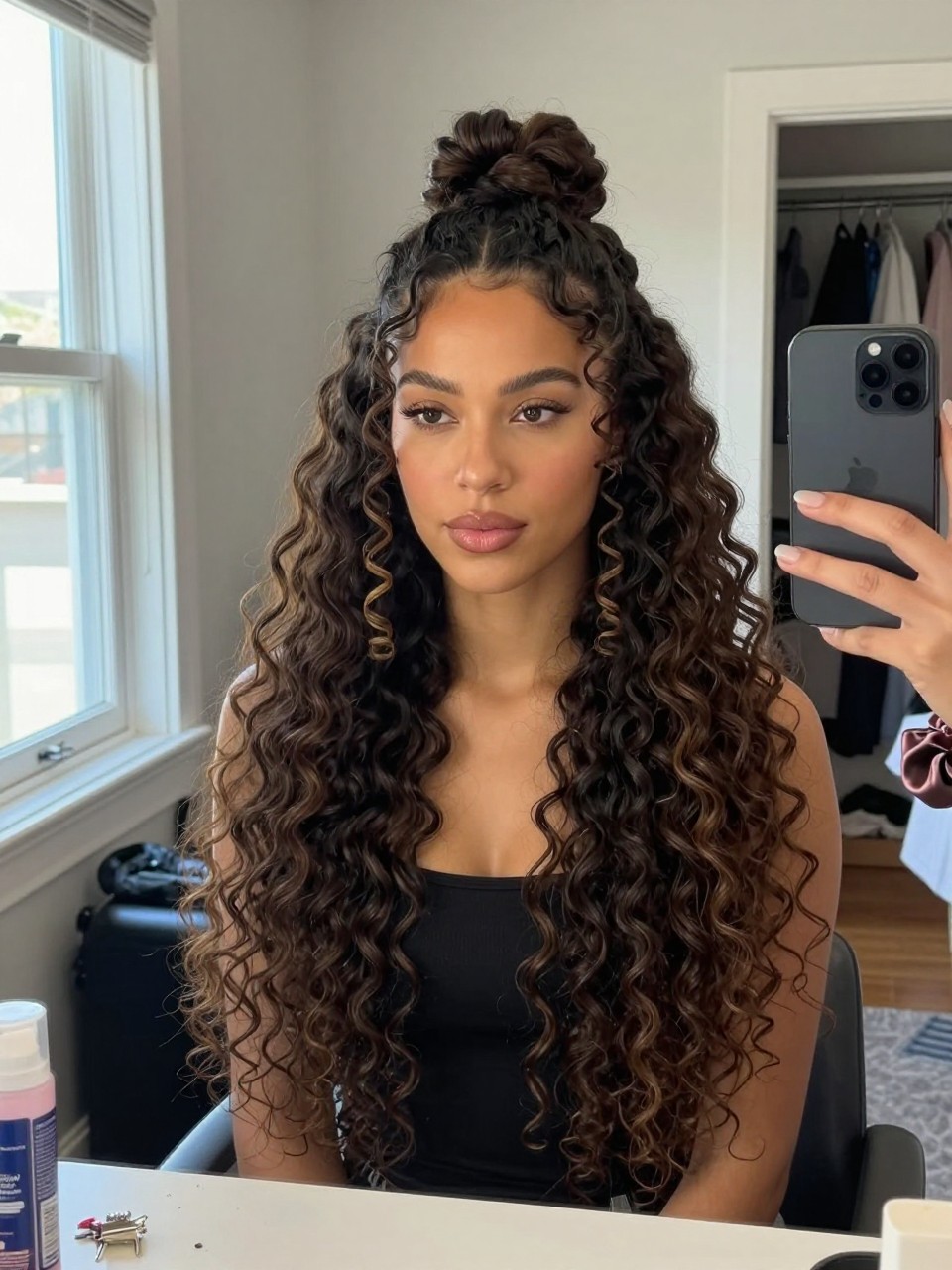 Image d'illustration : Photo d'une femme latine portant de longues boucles de type 4A en un demi-chignon avec une forme arrondie et douce au sommet de la tête, des boucles bien définies tombant en cascade en dessous, et deux mèches en spirale encadrant le visage, selfie pris sur le vif devant le miroir de sa chambre, lumière naturelle de la fenêtre, contenant un petit détail du quotidien comme des épingles à cheveux et un flacon de rafraîchisseur de boucles sur la commode, un chouchou en soie à son poignet et un placard légèrement ouvert derrière elle, moment réaliste de préparation, qualité photo iPhone de dernière génération.