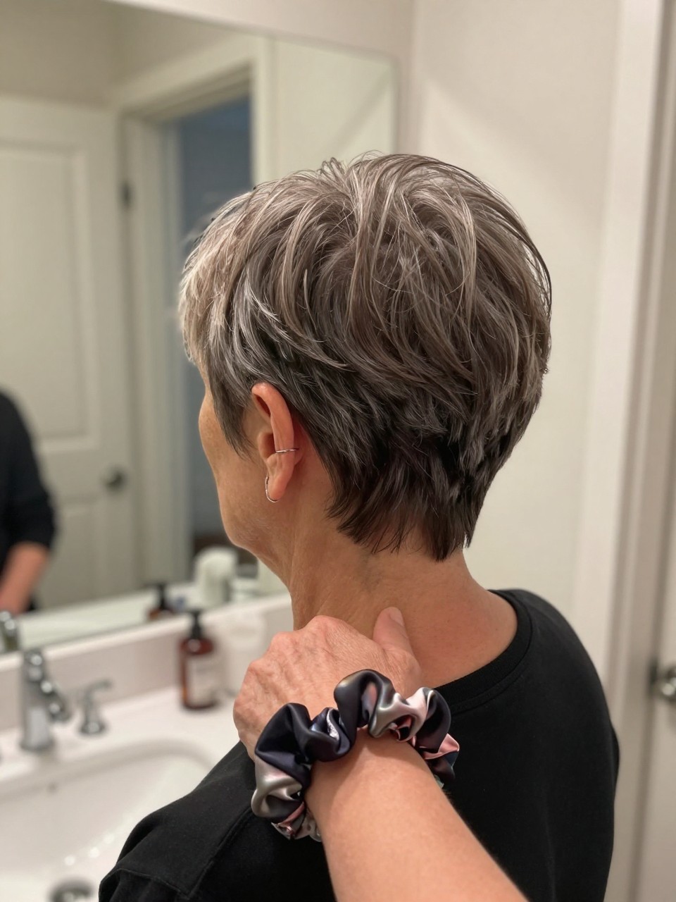 Photo d'une femme âgée portant une coupe pixie effilée à la nuque, vue de l'arrière de la tête montrant la ligne effilée nette au niveau de la nuque, le décor est un miroir de coiffeuse de salle de bain avec un éclairage intérieur doux, avec un chouchou en soie au poignet, qualité photo iPhone.
