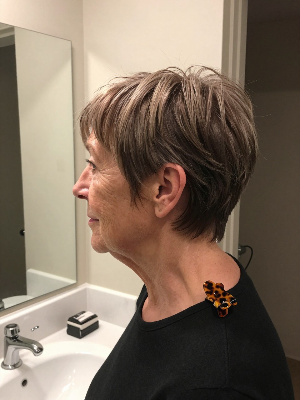 Photo d'une femme âgée portant une coupe pixie avec une frange balayée sur le côté, vue de profil montrant le balayage diagonal sur le front, le décor est un miroir de coiffeuse de salle de bain avec un éclairage intérieur doux, contenant une pince à cheveux en écaille de tortue sur son col, qualité photo iPhone.