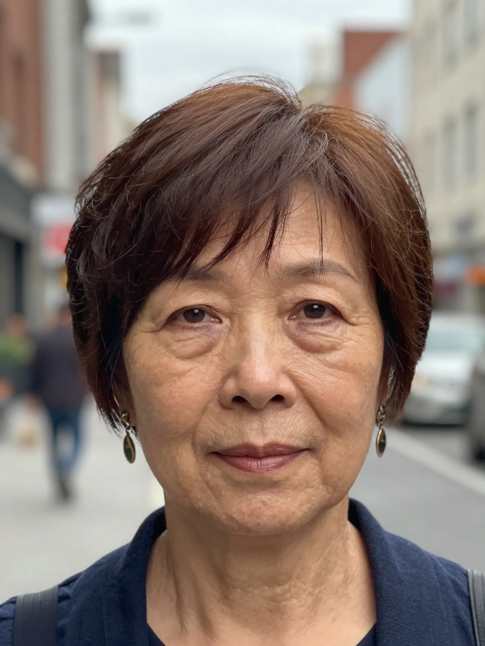 Photo d'une femme âgée portant une coupe pixie à frange souple, vue de face montrant une frange vaporeuse effleurant le front, le décor étant une rue de ville floue avec une douce lumière voilée, avec des boucles d'oreilles imposantes visibles, qualité photo iPhone.
