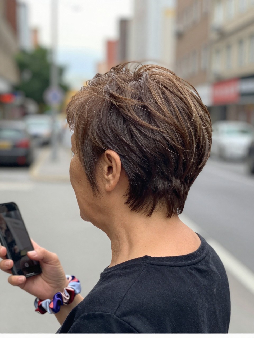 Photo d'une femme âgée portant une coupe pixie volumineuse à plusieurs niveaux, vue de l'arrière de la tête montrant le volume au niveau du sommet de la tête, le décor étant une rue de ville floue avec une douce lumière nuageuse, avec un chouchou en soie au poignet, qualité photo iPhone.