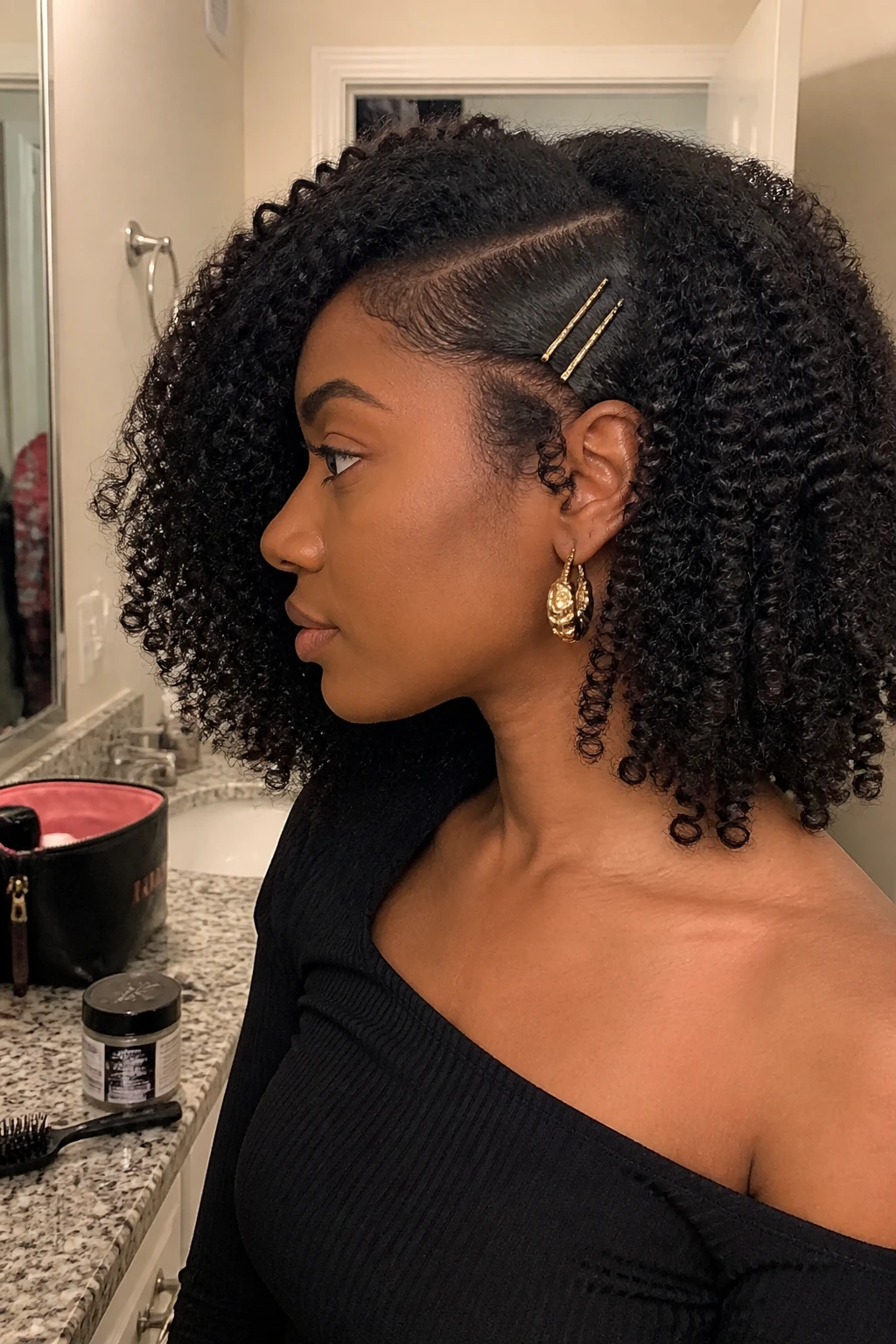 Image d'illustration : Photo d'une femme aux cheveux mi-longs bouclés de type 4A, avec une raie profonde sur le côté, une mèche soigneusement fixée au-dessus de l'oreille à l'aide de deux fines barrettes dorées, et des boucles bien définies balayant la joue opposée, vue de profil à côté d'un miroir de salle de bain, éclairage intérieur doux, avec un petit détail réaliste comme du gel pour les bords et une petite brosse sur le comptoir, une trousse de maquillage à proximité et des boucles d'oreilles imposantes qui dépassent, moment réaliste de préparation, qualité photo iPhone de dernière génération.