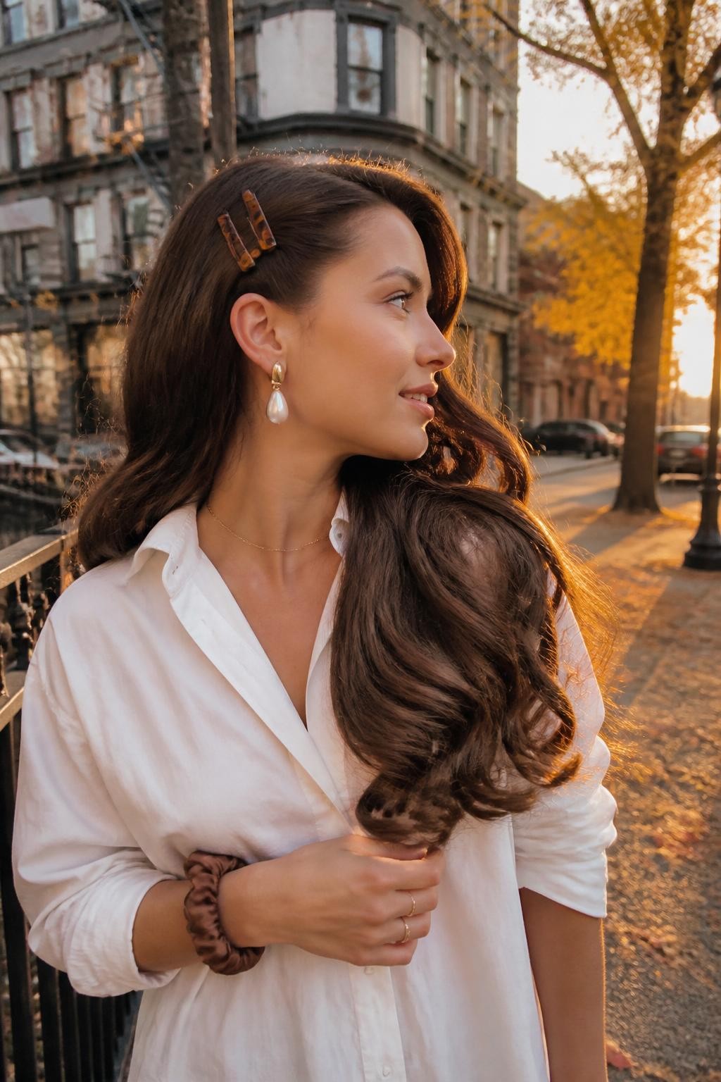 Photo d'une femme aux longs cheveux bouclés et brillants, avec une raie profonde sur le côté, une mèche relevée derrière l'oreille à l'aide de trois barrettes ambrées, vue de profil prise sur le vif dans une rue légèrement floue avec des feuilles dorées, une douce lueur de coucher de soleil, une boucle d'oreille remarquable visible du côté relevé et un chouchou en soie autour du poignet, mouvement réaliste des boucles dans la brise, qualité photo iPhone de dernière génération.
