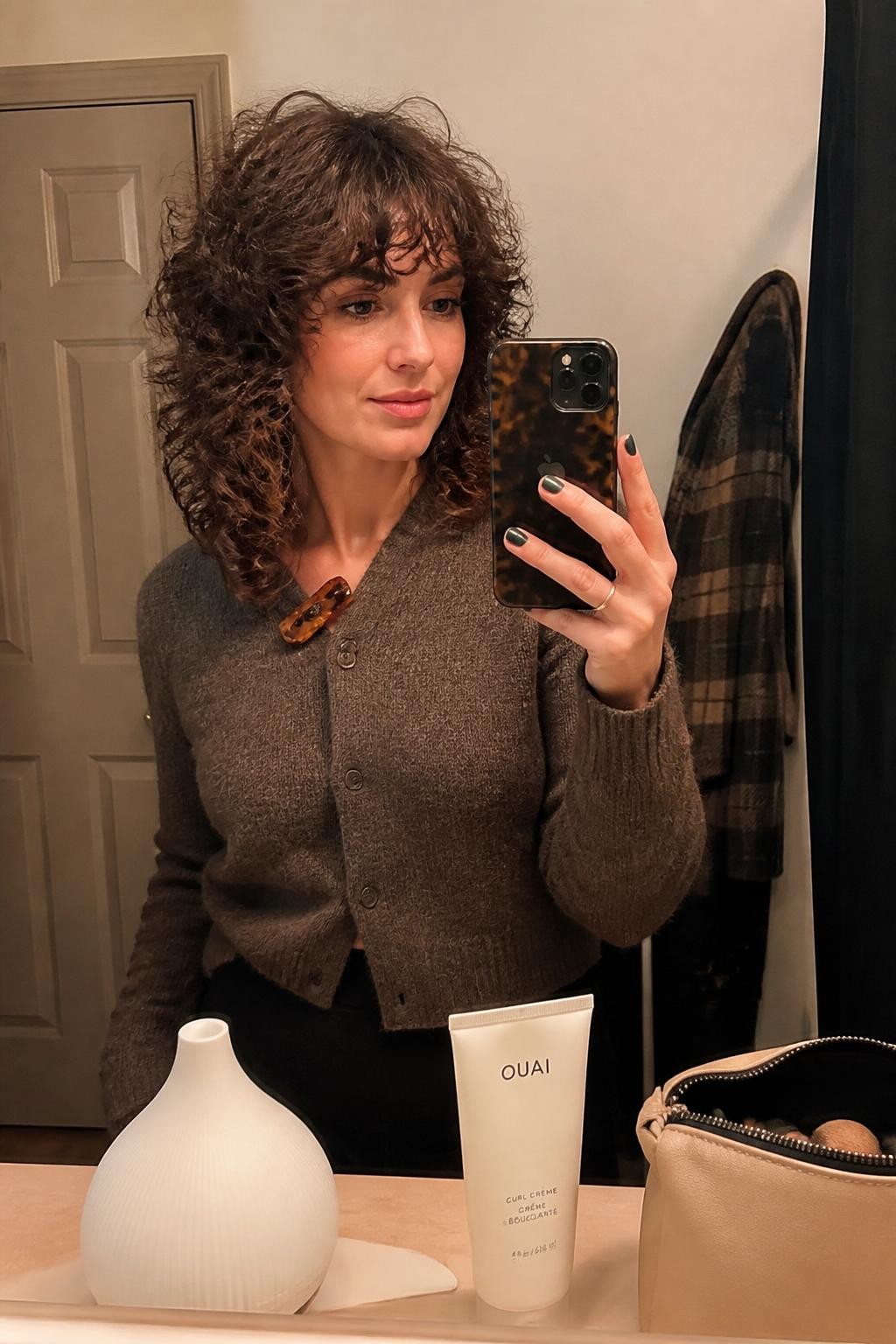 Photo d'une femme portant une coupe shag bouclée mi-longue avec des mèches courtes et aérées sur le dessus de la tête, une frange droite et des pointes légèrement effilées, selfie pris sur le vif dans un miroir de salle de bain chaleureux, éclairage intérieur doux, diffuseur et crème pour boucles posés sur le comptoir à côté d'une trousse de maquillage, pince à cheveux écaille de tortue attachée au col de son cardigan et un manteau à carreaux visible derrière elle, séparation des boucles et mèches rebelles réalistes, qualité photo iPhone de dernière génération.