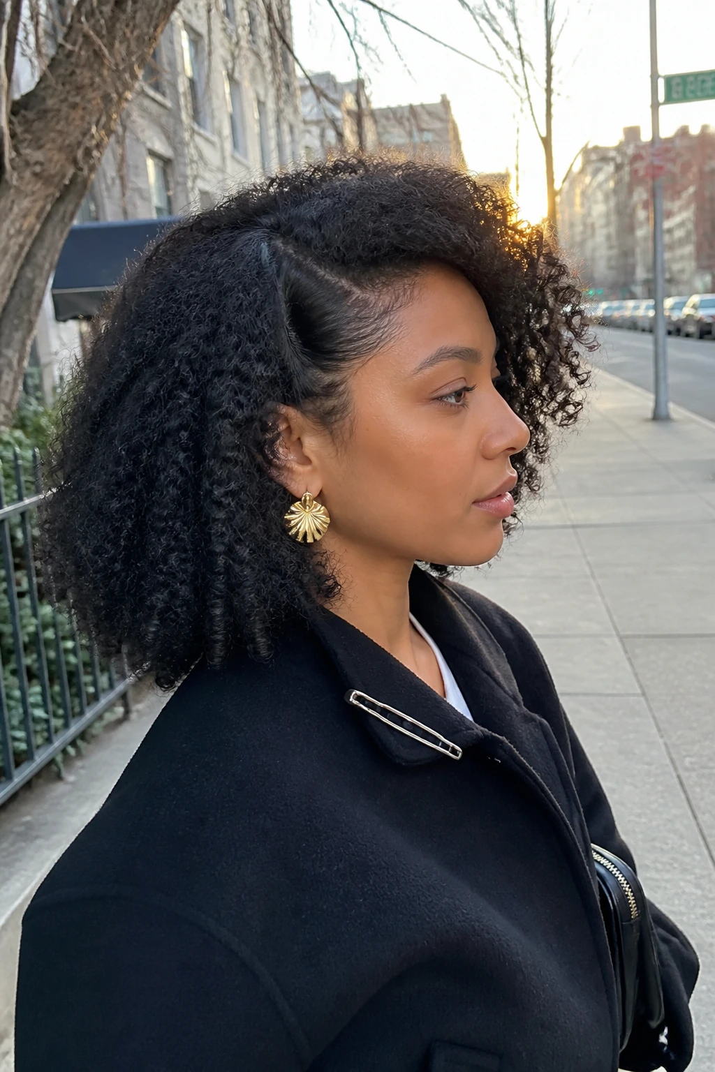 Image d'illustration : Photo d'une femme latine portant des boucles 4A étirées jusqu'aux épaules avec une raie profonde sur le côté, des boucles souples et allongées, des racines soulevées et un volume maximal balayé sur un côté, vue de profil sur fond de rue légèrement floue, lueur dorée de l'heure dorée, contenant un petit détail du quotidien comme des boucles d'oreilles imposantes, une fine pince à cheveux attachée au col de sa veste et un petit sac à main près de son coude, photographie réaliste prise sur le vif, qualité photo iPhone de dernière génération.