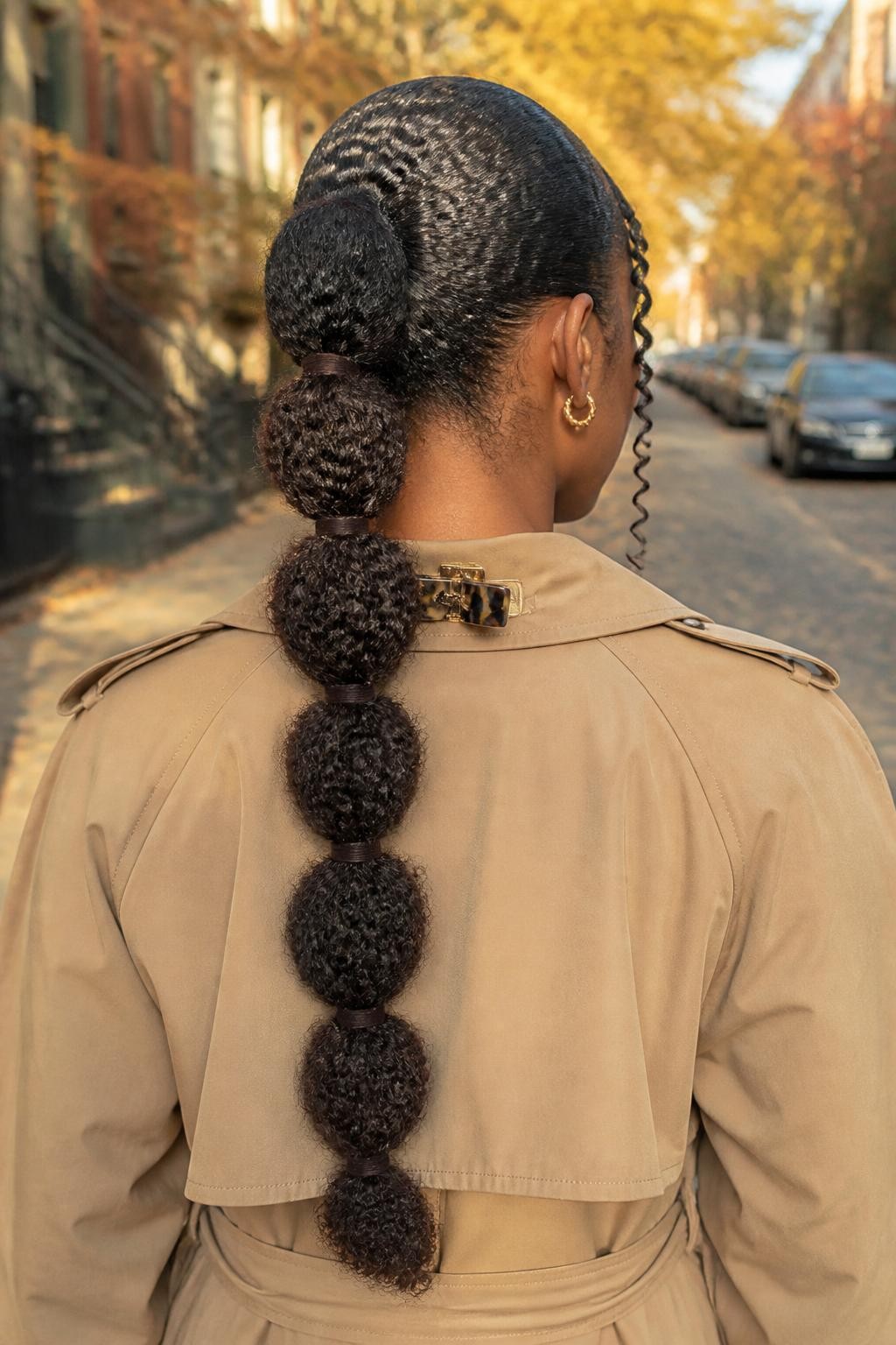 Photo d'une femme noire portant une longue queue de cheval texturée à bulles avec des sections bouclées arrondies séparées par d'étroits liens en velours chocolat, des bords souples et deux mèches en spirale encadrant son visage, vue de l'arrière de la tête sur fond de rue d'automne légèrement floue, lumière dorée, pince à cheveux attachée au col de son trench-coat et petites boucles d'oreilles en or visibles, texture de boucle naturelle réaliste, qualité photo iPhone de dernière génération.
