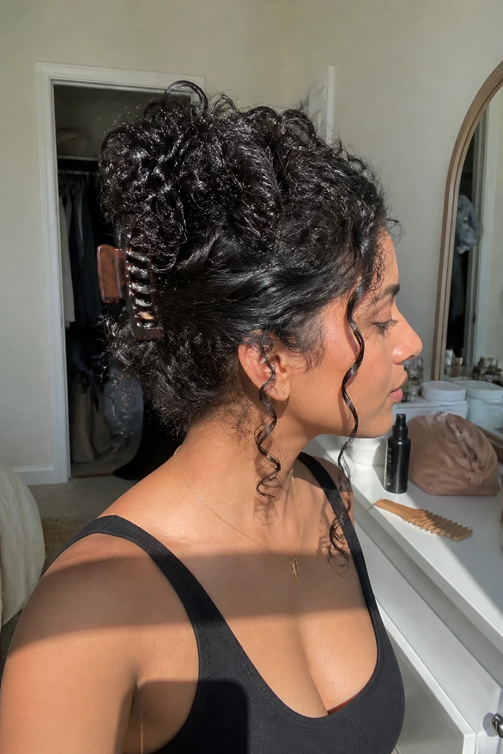 Image d'illustration : Photo d'une femme sud-asiatique portant de longues boucles de type 4A relevées en chignon avec une pince à cheveux, les pointes des boucles denses retombant doucement sur une grande pince écaille de tortue, et deux boucles encadrant le visage laissées libres, vue de profil dans une chambre baignée de soleil, lumière naturelle de la fenêtre, contenant un petit détail du quotidien tel qu'un peigne à dents larges et un flacon de spray pour boucles sur la commode, une trousse de maquillage à proximité et un placard légèrement ouvert derrière elle, moment réaliste de préparation, qualité photo iPhone de dernière génération.
