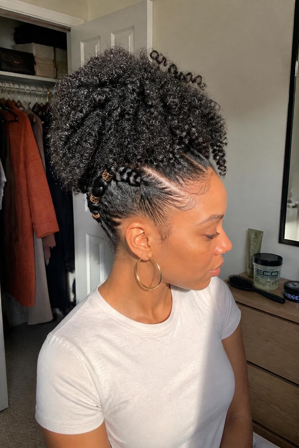 Photo d'une femme noire portant une coiffure volumineuse et sculptée avec des boucles naturelles souples, deux torsades latérales soignées menant vers le sommet de la tête, des bords légèrement travaillés et de petits bracelets en bronze placés le long des torsades, vue de profil dans une chambre baignée de soleil, lumière naturelle chaude provenant d'une fenêtre, brosse à bords et gel pour boucles sur la commode, cardigan rouille suspendu dans un placard légèrement ouvert et créoles dorées qui apparaissent, texture naturelle réaliste, qualité photo iPhone de dernière génération.