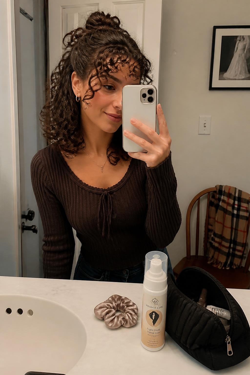 Photo d'une femme aux cheveux bouclés mi-longs, coiffés en un demi-chignon décontracté avec une frange bouclée lâche et des longueurs légèrement définies, selfie pris sur le vif dans un miroir de salle de bain, éclairage doux et chaud, chouchou en soie et mousse à boucles posés à côté d'une trousse de maquillage, petites boucles d'oreilles créoles visibles et une écharpe à carreaux drapée sur une chaise, un moment de coiffure réaliste du quotidien, qualité photo iPhone de dernière génération.