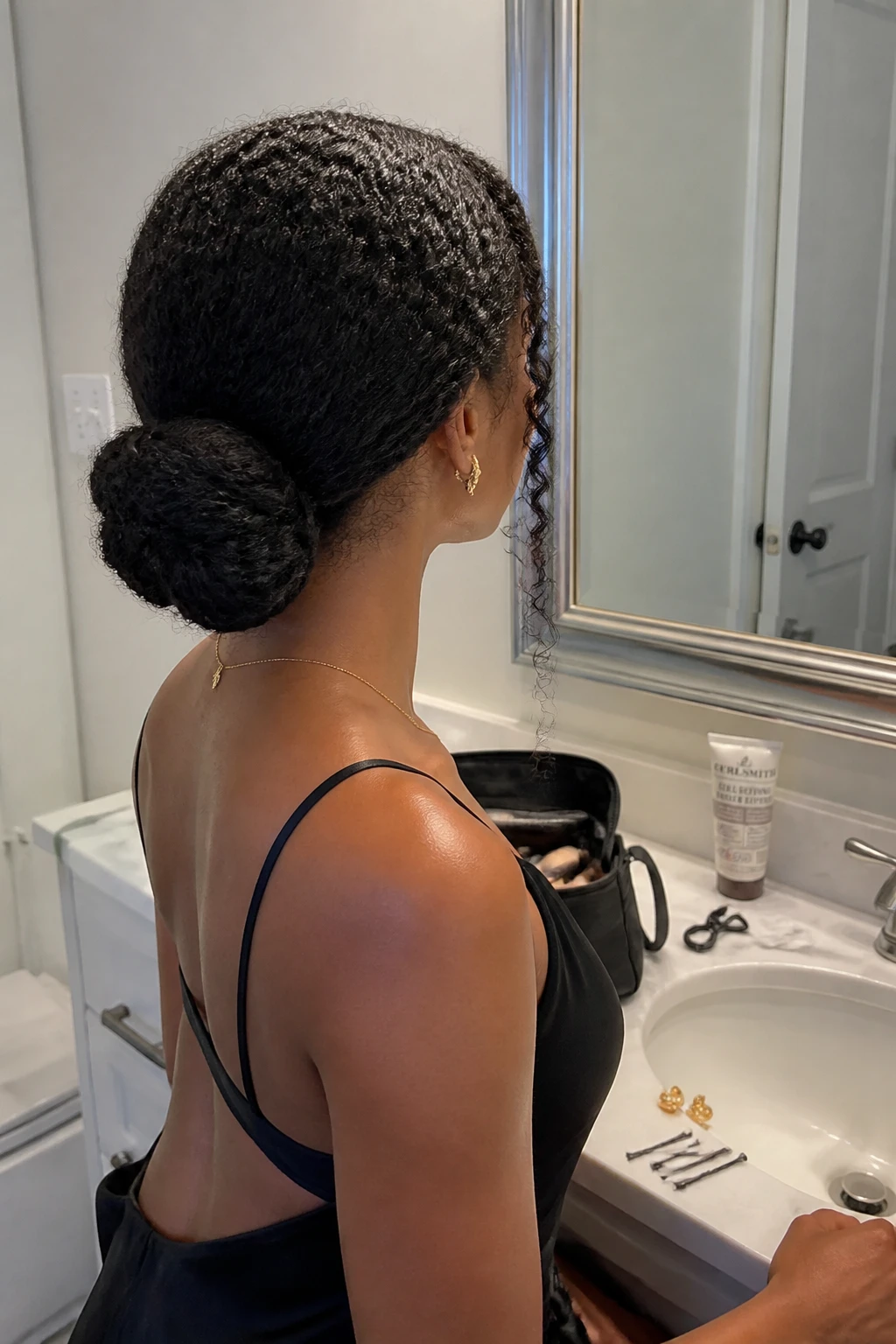 Image d'illustration : Photo d'une femme portant un chignon bas torsadé, réalisé à partir de boucles denses de type 4A avec une raie au milieu nette, un volume souple sur le dessus de la tête et deux mèches en spirale bien définies encadrant le visage, vue de trois quarts arrière à côté d'un miroir de salle de bain, éclairage intérieur doux, contenant un petit détail réaliste comme des épingles à cheveux à côté d'une trousse de maquillage ouverte, de la crème pour boucles sur le comptoir et de petites boucles d'oreilles en or posées près du lavabo, moment réaliste de préparation formelle, qualité photo iPhone de dernière génération.